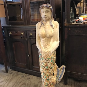 May include: A weathered, hand-carved wooden mermaid statue. The mermaid has blonde hair with blue accents, a nude torso, and a colorful, scaled tail in shades of green, blue, and gold. The statue is mounted on a blue base and shows signs of age.