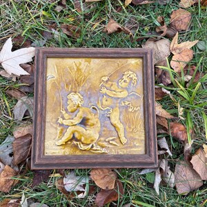 May include: A framed, square relief sculpture featuring two cherubic figures in a pastoral scene. The figures are rendered in a golden hue, set against a textured background. The frame is a dark brown color.