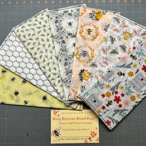 May include: A collection of six fabric bread bags arranged in a fan shape, featuring various bee, honeycomb, and floral patterns in yellow, white, and orange. A rectangular card is placed below, labeled Bizzy Beezwax Bread Bags with text.