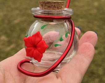 May include: A small glass hummingbird feeder with a red flower design and a cork stopper. The feeder is attached to a red metal wire hanger.