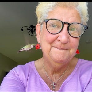 May include: A person with short blonde hair wearing black-rimmed glasses and a light purple long-sleeved shirt. They are wearing red flower-shaped earrings, with a small hummingbird hovering near the left earring, and a silver cross necklace.