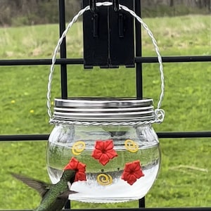 May include: A glass hummingbird feeder with a metal wire handle hangs from a black metal fence. The feeder is filled with clear liquid and decorated with red flower shapes and yellow swirls. A hummingbird is drinking from the feeder.