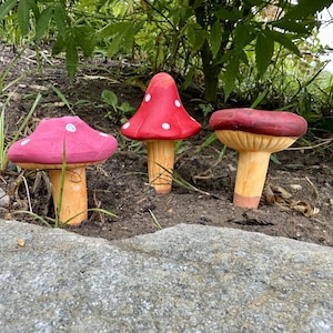 May include: Three decorative mushroom figurines with red, pink, and maroon caps and tan stems. The red mushroom has white polka dots. The mushrooms are placed in a garden setting with green foliage in the background.