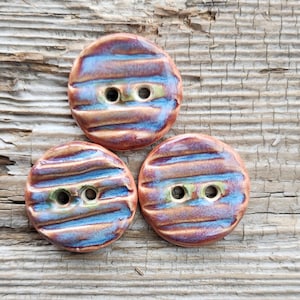 May include: Three handmade ceramic buttons with a glossy finish. The buttons are round with two holes and feature a striped pattern in shades of brown, blue, and green. The buttons are arranged on a weathered wooden surface.
