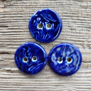 May include: Three round, glossy, cobalt blue ceramic buttons with two holes each. The buttons have a textured, swirled design in a lighter shade of blue. The buttons are arranged on a weathered, light brown wooden surface.