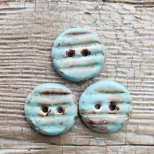 May include: Three handmade ceramic buttons with a light blue glaze and brown horizontal stripes. Each button has two holes for sewing. The buttons are round and have a textured surface, perfect for adding a unique touch to any sewing project.