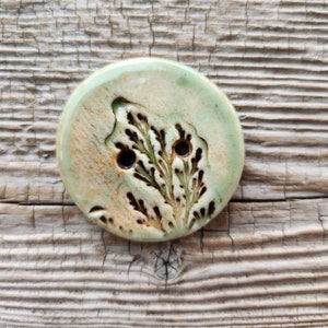May include: A round ceramic button with a green glaze and a brown fern design. The button has two holes for sewing.