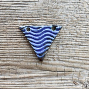 May include: A triangular ceramic bead with a wavy blue pattern. The bead has two holes for stringing and is set against a weathered, light brown wooden surface. The bead's edges are dark blue.