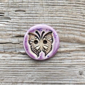 May include: A purple ceramic button with a detailed butterfly design. The butterfly is in shades of brown and white.