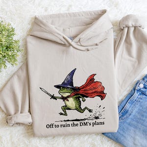 May include: A beige hooded sweatshirt featuring a graphic of a green frog wearing a purple wizard hat and a red cape, holding a sword. The text below the frog reads, Off to ruin the DM's plans. The sweatshirt has long sleeves and a drawstring hood.