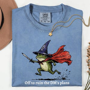 May include: A blue t-shirt featuring a graphic of a green frog wearing a purple wizard hat and a red cape, holding a sword. The text below the frog reads, Off to ruin the DM's plans. The shirt has a Comfort Colors label showing size XL.