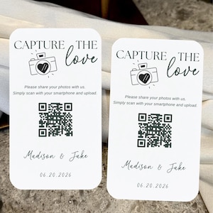 May include: Two white wedding cards with rounded corners. The cards have the text "CAPTURE THE love" above a camera icon with a heart. Below is a QR code, the names "Madison & Jake", and the date 06.20.2026.