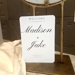 以下が含まれることがあります： エレガントな筆記体で「Madison + Jake」の名前が書かれた白いウェディングウェルカムサイン。サインには結婚式の日付と場所が記載されており、金色の金属製のスタンドに表示されています。サインには「Welcome to the wedding of」と書かれています。