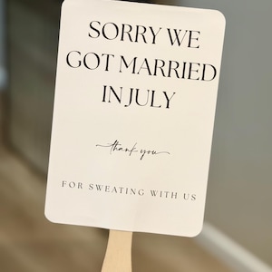 Può includere: Un ventaglio da matrimonio rettangolare bianco con il testo nero "SORRY WE GOT MARRIED IN JULY", con "thank you" in corsivo e "FOR SWEATING WITH US" sotto. Ha un manico di legno.