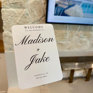 以下が含まれることがあります： 「WELCOME TO THE WEDDING OF Madison + Jake」と書かれた白いウェディングファン。ファンには場所と日付も含まれています：Nashville, TN 08.09.2025。ファンは木の柄で持たれています。