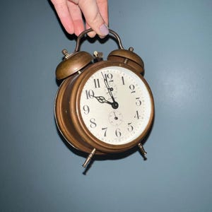 May include: A vintage brass alarm clock with a cream-colored face and black numerals. The clock has two bells on top and a handle for carrying. The clock is held by a hand against a blue-gray background.