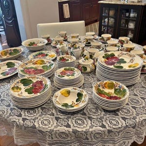 May include: A large collection of hand-painted ceramic dinnerware, including plates, bowls, cups, saucers, and serving dishes. The floral design features red, yellow, and green fruits and vegetables. The set is arranged on a round table covered with a white lace tablecloth.