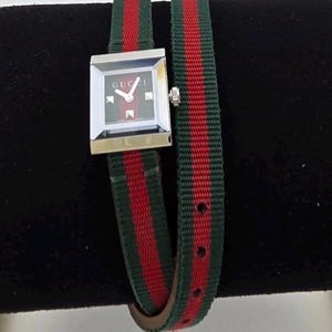 Könnte beinhalten: Gucci-Uhr mit quadratischem silbernem Zifferblatt und grün-rot gestreiftem Armband. Das Zifferblatt ist dunkel mit silbernen Details und dem Gucci-Logo. Das Armband wickelt sich zweimal um das Handgelenk.