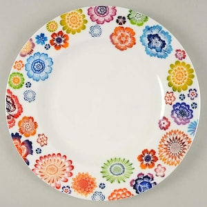 Puede incluir: Plato de cerámica blanco con un diseño floral vibrante. El plato presenta un borde de flores coloridas en tonos de rojo, naranja, amarillo, verde, azul y morado. Las flores varían en tamaño y estilo, creando un aspecto alegre y decorativo.