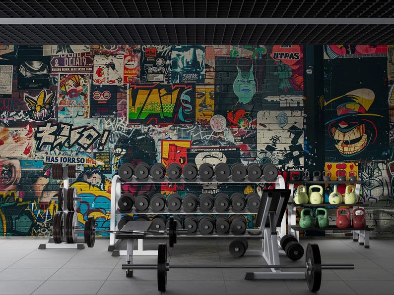 May include: A gym interior with a graffiti art brick wall backdrop. Weightlifting equipment includes barbells, dumbbells, and kettlebells. The room has a modern, industrial aesthetic with a black ceiling.
