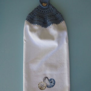 May include: A white kitchen towel with a blue crocheted top and a button. The towel has a design of three seashells in the bottom right corner.