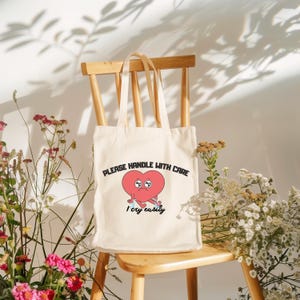 Pode incluir: Saco tote creme com o texto "PLEASE HANDLE WITH CARE" e "I cry easily". O saco apresenta um coração de desenho animado com olhos tristes e braços. O saco está numa cadeira de madeira, rodeado por flores.