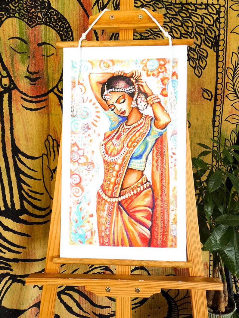 May include: An artwork depicting a woman in traditional Indian attire, adorned with jewelry and a headpiece. The artwork is displayed on a wooden easel, with a backdrop of patterned fabric. The woman is wearing a blue top and an orange sari.