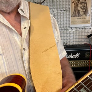 May include: A wide, light tan leather guitar strap with a natural, textured surface and visible grain, worn over a white button-down shirt. The strap is attached to a cherry-red electric guitar with a yellow-trimmed body and a wooden fretboard.