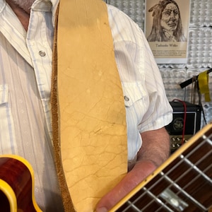 May include: A wide, light tan leather guitar strap with a natural, textured surface rests over a white pinstriped shirt. The strap features a raw, suede-like underside and is attached to a cherry-red electric guitar with a yellow binding.