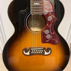 May include: Acoustic 12-string guitar with a sunburst finish, transitioning from black to brown and yellow. The guitar features a decorative pickguard with floral designs and a bridge with 12 pins. The fretboard has inlaid markers.