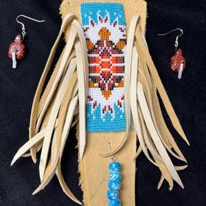 May include: A tan leather necklace featuring a central rectangular panel with a beaded turtle design in blue, white, orange, and brown, surrounded by long leather fringe. The set includes matching earrings with brown turtle-shaped charms.