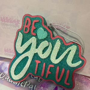 May include: A teal and glittery ornament with the words "BE you TIFUL" in white and red. The ornament has a pink border and a small teal heart. A white string is attached for hanging.