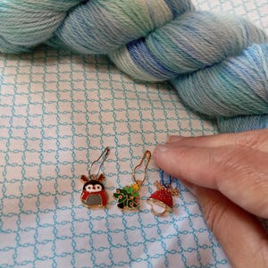 May include: Three gold-colored stitch markers with Christmas-themed designs. The markers feature a penguin, a Christmas tree, and a red and white bell.