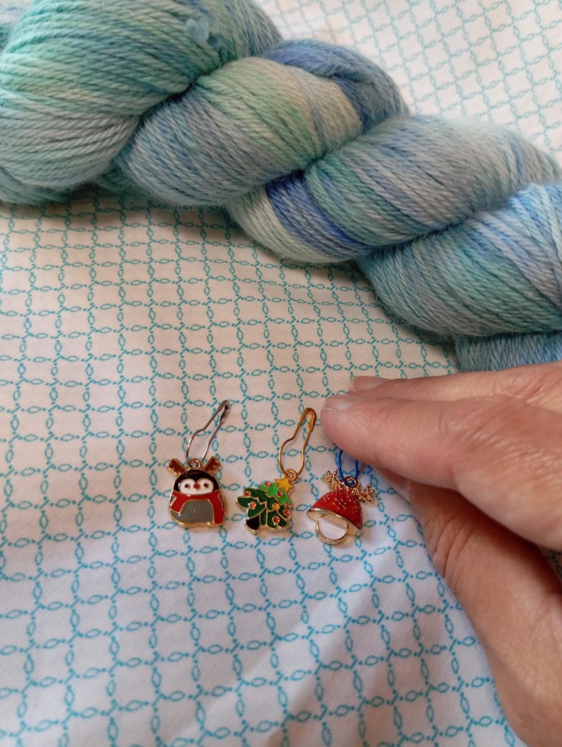 May include: Three gold-colored stitch markers with Christmas-themed designs. The markers feature a penguin, a Christmas tree, and a red and white bell.