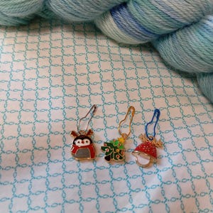 May include: Four colorful Christmas themed stitch markers. The markers are shaped like a penguin, a Christmas tree, and a bell. The markers are made of metal and have a gold finish.