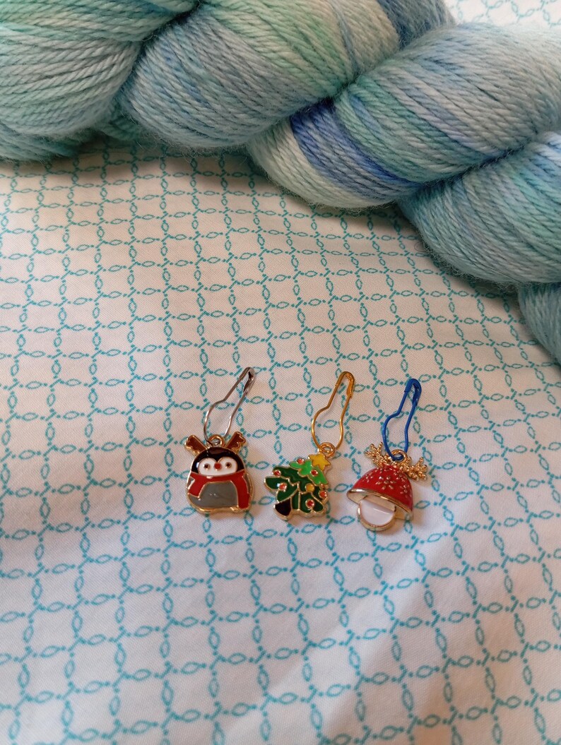 May include: Four colorful Christmas themed stitch markers. The markers are shaped like a penguin, a Christmas tree, and a bell. The markers are made of metal and have a gold finish.