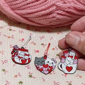 May include: Three cat-themed stitch markers with red and white hearts. The first marker is a glass jar with a red ribbon and the text "Filled with Love". The second marker is a heart shape with two cats inside. The third marker is a white mug with a red heart and a cat inside.