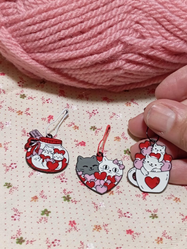 May include: Three cat-themed stitch markers with red and white hearts. The first marker is a glass jar with a red ribbon and the text "Filled with Love". The second marker is a heart shape with two cats inside. The third marker is a white mug with a red heart and a cat inside.