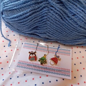 May include: Three Christmas-themed stitch markers with a reindeer, a Christmas tree, and a bell. The markers are on a white card with pink and blue stripes and the text "www.stitchesmarked.etsy.com".