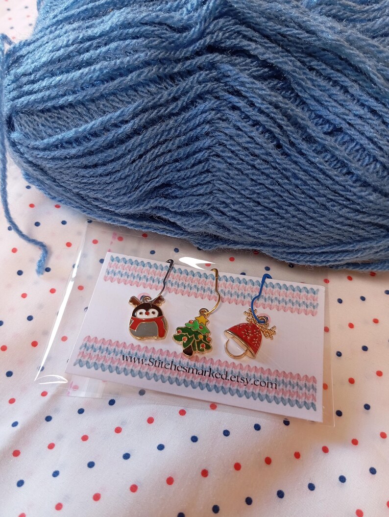May include: Three Christmas-themed stitch markers with a reindeer, a Christmas tree, and a bell. The markers are on a white card with pink and blue stripes and the text "www.stitchesmarked.etsy.com".