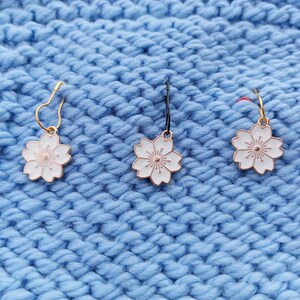 May include: Three white flower-shaped stitch markers with gold and black wire loops. The markers are on a blue knitted fabric background.