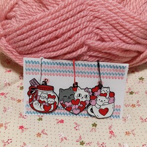 May include: Three cat-themed stitch markers with red hearts. The first marker is a glass jar with a label that says "Full of Love". The second marker is a heart with a gray cat inside. The third marker is a white mug with a cat inside.