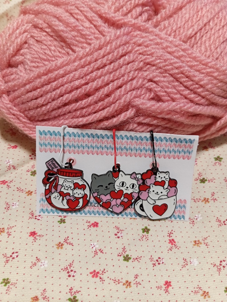 May include: Three cat-themed stitch markers with red hearts. The first marker is a glass jar with a label that says "Full of Love". The second marker is a heart with a gray cat inside. The third marker is a white mug with a cat inside.