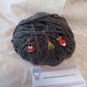 May include: Three gold stitch markers with different Christmas-themed charms: a penguin, a Christmas tree, and a red and white mushroom. The markers are on a ball of gray yarn. A business card with the text "www.Stitchesmarked.etsy.com" is in the foreground.