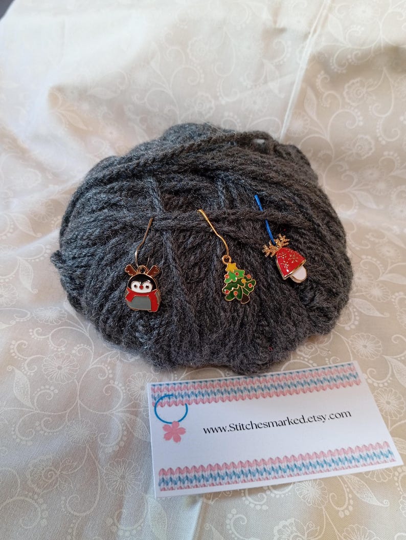 May include: Three gold stitch markers with different Christmas-themed charms: a penguin, a Christmas tree, and a red and white mushroom. The markers are on a ball of gray yarn. A business card with the text "www.Stitchesmarked.etsy.com" is in the foreground.