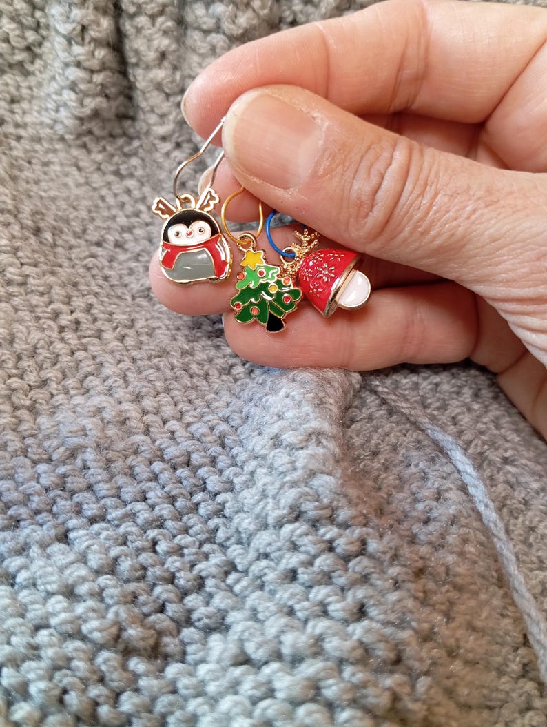 May include: Three Christmas themed stitch markers with gold rings. The markers feature a penguin, a Christmas tree, and a bell with a snowflake design.