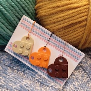 Può includere: Tre orecchini a forma di cuore realizzati con mattoncini giocattolo ad incastro beige, arancioni e marroni. Gli orecchini sono su una carta bianca con un bordo rosa e blu, su uno sfondo di filato verde acqua, giallo senape e azzurro.