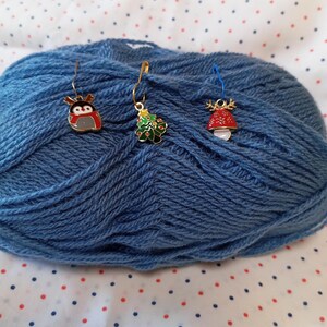 May include: Three gold metal stitch markers with different Christmas-themed charms. The charms are a penguin, a Christmas tree, and a mushroom.