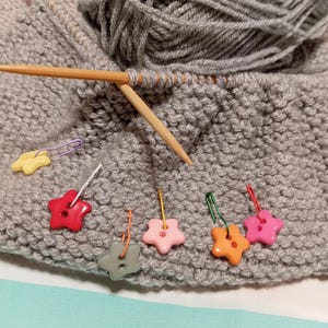 Op de afbeelding: Een close-up van een breiproject met grijs garen, houten breinaalden en stervormige steekmarkeerders in verschillende kleuren. Het garen is in een bol en wordt ook verwerkt in een gebreid stuk.