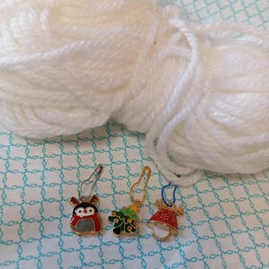 May include: Three Christmas themed stitch markers with a penguin, a Christmas tree, and a bell. The markers are gold with colorful enamel details. The markers are on a white background.